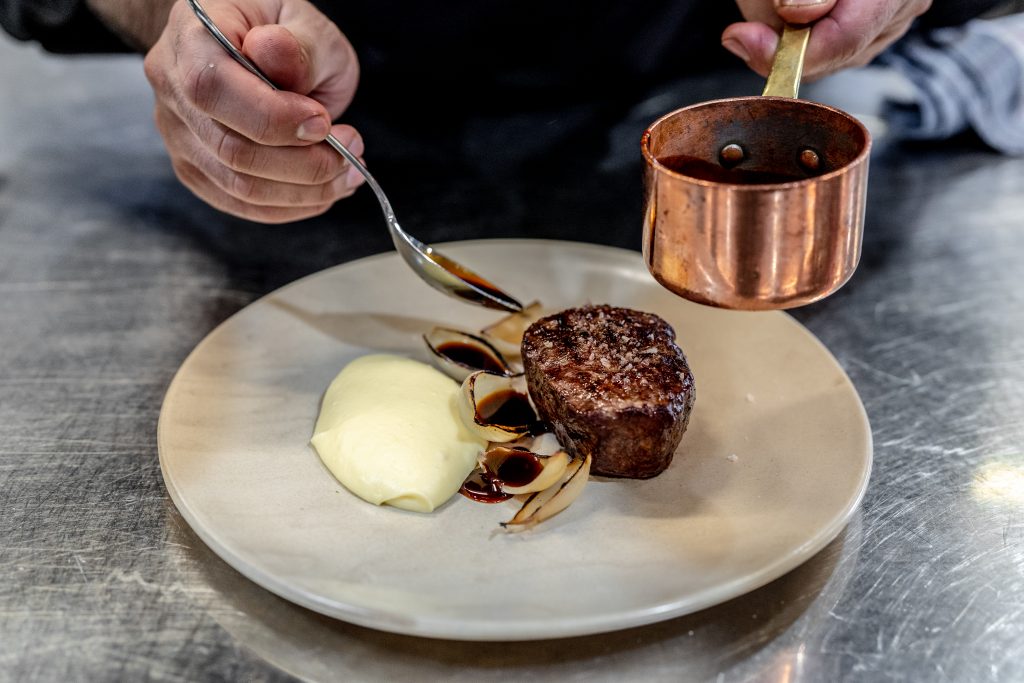 A chef dresses a steak on a plate with a side of mash potato