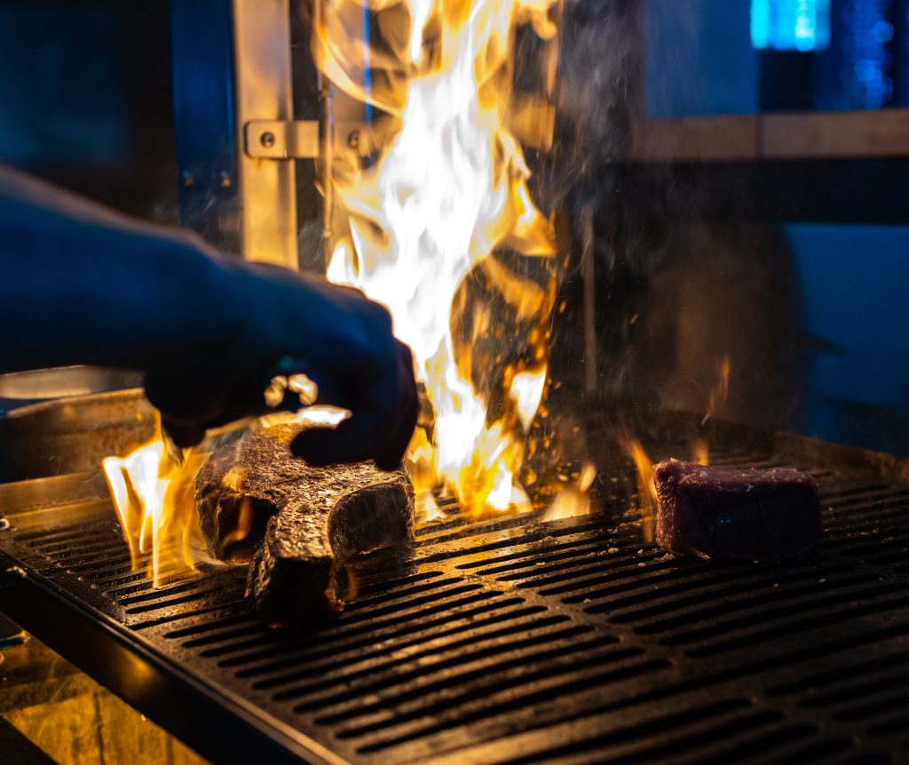 A chef tends to a steak on a flaming grill